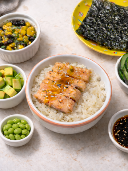 A salmon and rice bowl for toddlers is shown surrounded by various toppings: broccoli and corn salad, seaweed snacks, pickles, soy dressing, edamame beans, and avocado cubes.