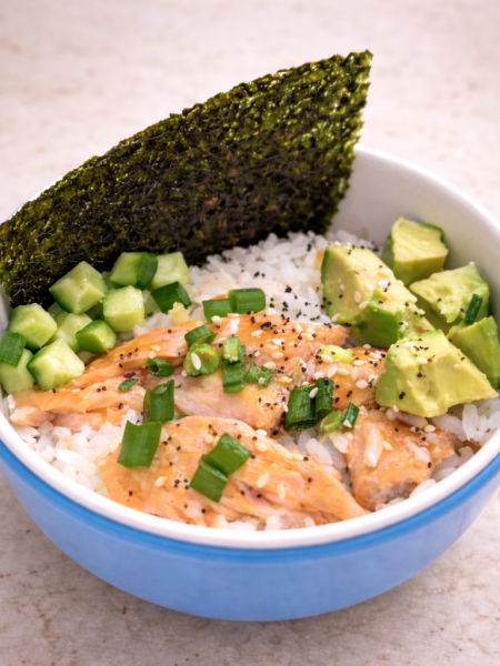 A salmon rice bowl for toddlers is shown stuffed with avocado cubes, seaweed strips, and diced cucumber.