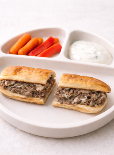 A toddler philly cheesesteak is shown sliced in half and plated with carrots and ranch.