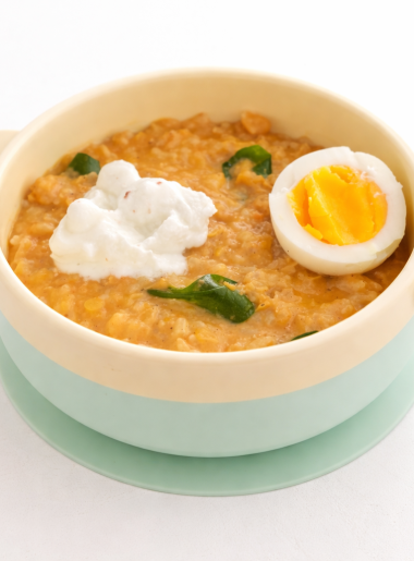 A child's bowl filled with red lentil and rice curry and topped with yogurt, spinach, and half a hard-boiled egg.