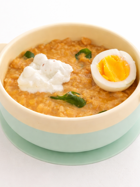 A child's bowl filled with red lentil and rice curry and topped with yogurt, spinach, and half a hard-boiled egg.