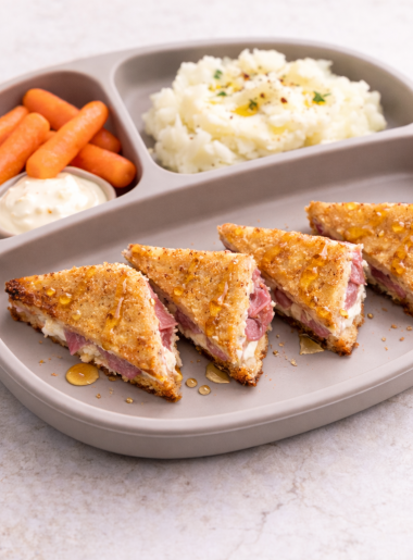 A toddler grilled sandwich stuffed with meat and cheeseis shown drizzled in honey and cut into triangles Spanish "bikini" style; mashed potatoes and cut carrots side the sandwich.