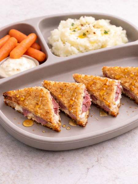 A toddler grilled sandwich stuffed with meat and cheeseis shown drizzled in honey and cut into triangles Spanish "bikini" style; mashed potatoes and cut carrots side the sandwich.