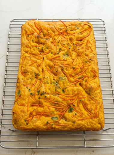 A loaf of carrot focaccia for kids, topped with chives and carrot strands, is shown on a cooling rack.