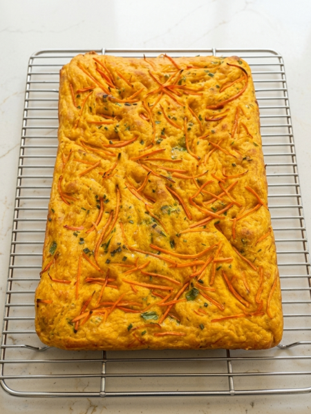 A loaf of carrot focaccia for kids, topped with chives and carrot strands, is shown on a cooling rack.