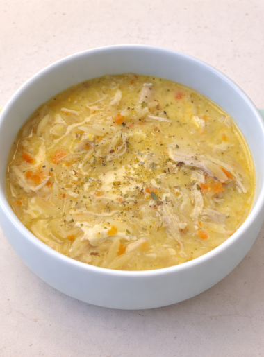 A small blue bowl is shown filled with a lemon chicken orzo soup, with visible shreds of poultry, carrot pieces, and oregano.