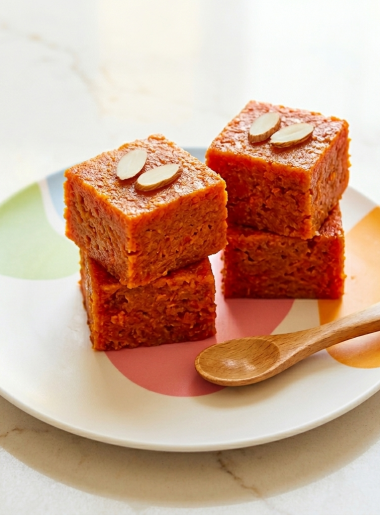 Small stacks of sweet carrot bites for toddlers (carrot halwa) are shown topped with slivered almonds and shown on a plate, with a small spoon in front.