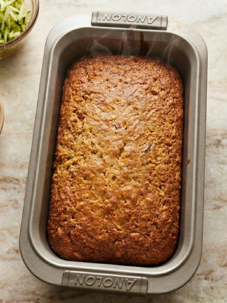 A loaf pan holding honey nut zucchini bread for kids; bowls of grated zucchini and pecans are shown at its side.