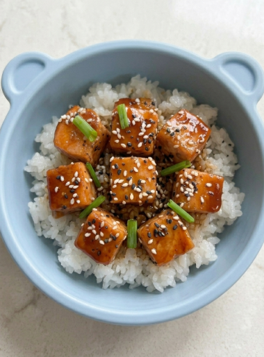 A small dinner bowl holds glazed teriyaki salmon for kids atop rice, with sesame seeds and chives strewn atop the fish.