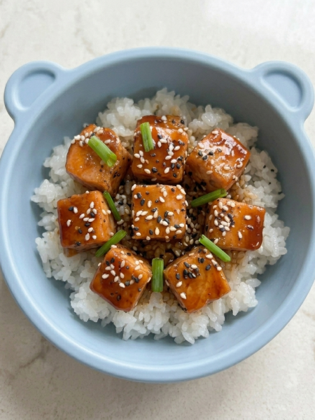 A small dinner bowl holds glazed teriyaki salmon for kids atop rice, with sesame seeds and chives strewn atop the fish.