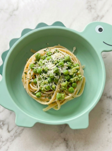 A bowl of mashed pea pasta for kids is shown, with peas and grated cheese strewn over the top of spaghetti noodles.