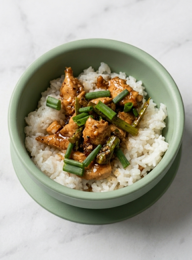 A bowl of toddler three cups chicken, with nubbins of chicken and sticks of asparagus strewn atop white rice.