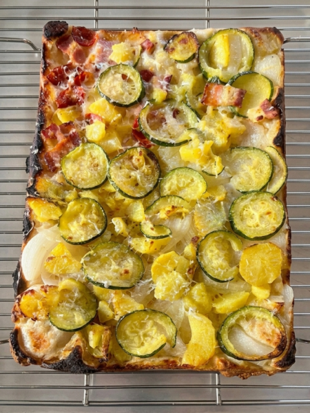 An upside down pizza for kid is covered in toppings like potato, zucchini, cherry tomatoes, and bacon slices.