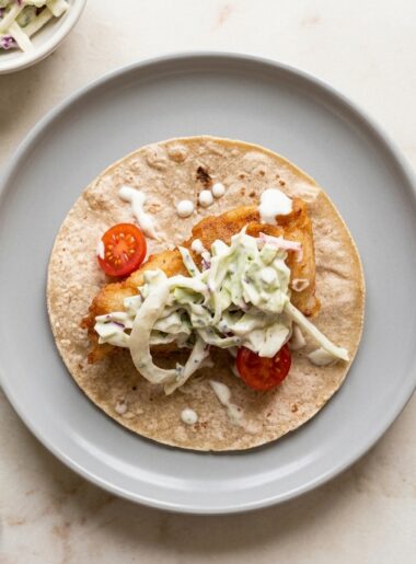 Overhead shot of a corn tortilla holding a plank of fried fish, which is topped with creamy slaw, dots of sauce, and a few tomato slices. Fish tacos for kids