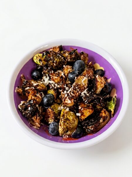 A small bowl of brussels sprouts for kids with crisp ridges is shown; blueberries and grated parmesan are also visible atop the sprouts.